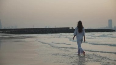 Arka planda Dubai gökdelenlerinin silueti ile suda yürüyen esmer kadın var.