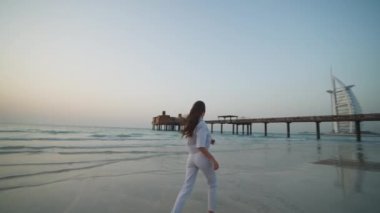 Dubai plajında çıplak ayakla koşan genç bir kadın ve arka planda Burj Al Arab var.