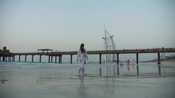 Niski kąt widzenia młodej brunetki w eleganckich ubraniach spacerujących po piaszczystej plaży w kierunku hotelu Burj Al Arab w Dubaju — Wideo stockowe