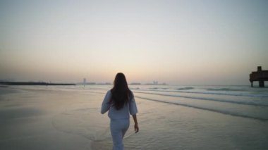 Arka planda zarif elbiseli bir kadın kumsalda suda yürüyor ve arka planda Dubai şehri manzarası var.