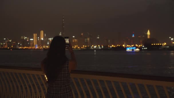 夜にドバイのスカイラインの写真を撮る女性。彼女の携帯電話でドバイの高層ビルの写真を撮るブルネットの女性 — ストック動画