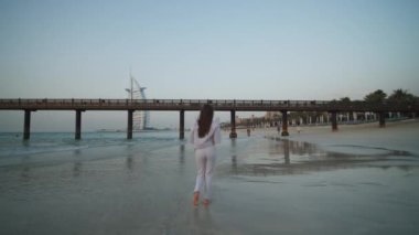 Arka planda Burj Al Arap oteliyle Dubai 'de plajda koşan genç beyaz kadın.