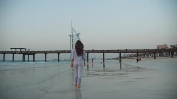Вид на привлекательную молодую брюнетку с длинными волосами, идущую по песчаному пляжу в Дубае в направлении отеля Burj Al Arab — стоковое видео