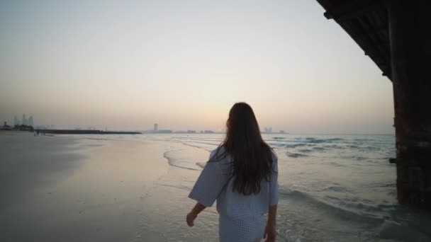 Junge Frau spaziert unter Holzsteg am Sandstrand mit Dubais Skyline im Hintergrund — Stockvideo
