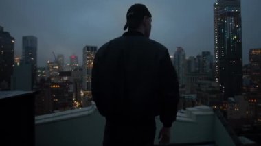 Geceleri New York 'ta bir gökdelenin çatısında duran bir adam. Arka planda şapkalı ve ceketli bir adam çatıdan New York şehrine bakıyor.