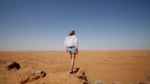 Mujer joven de pie en la cima de una colina rocosa con vistas a un vasto desierto. Turista explorando un desierto cerca de Dubai en un día soleado — Vídeo de stock