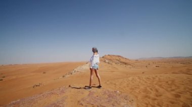 Beyaz gömlekli kadın kum tepeciğinin kenarında durup çöle bakıyor. Başında atkıyla çölde dikilen bir turist.