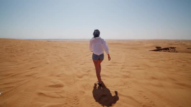 Seguindo uma jovem caminhando no deserto. Visão traseira da jovem mulher de camisa e jeans andando sobre dunas de areia — Vídeo de Stock