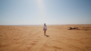 Dubai yakınlarındaki çölde yürüyen güneş gözlüklü ve eşarplı genç bir kadının ön görüntüsü.