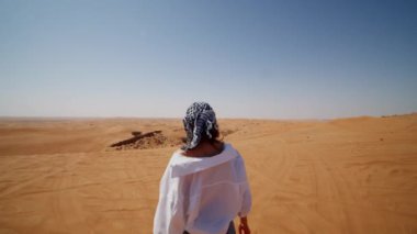 Çölde kum tepelerinde yürüyen atkı ve güneş gözlüklü beyaz bir kadın.