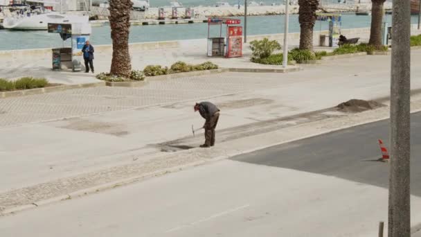 Yol asfalt katmanını yenilemeden önce yüzeyi tamamlamak için kazma kullanan sokak işçisinin yüksek görüntüsü. Portekiz, Lagos sokaklarında fiziksel çalışma. — Stok video