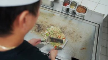Sıcak tabakta sebzeli erişteye fasulye filizi, mantar ve soya sosu ekleyen bir aşçı. Restoranın mutfağında sebzeli erişte karıştıran profesyonel aşçı manzarası.