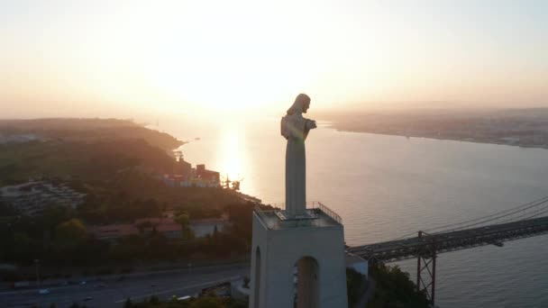 Atemberaubende Umlaufbahn des Heiligtums von Christus König Statue auf dem Hügel im Abendlicht mit Enthüllung der Ponte 25 de Abril rote Brücke und Lissabon Stadtzentrum über das Meer — Stockvideo