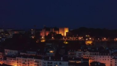 Şehrin yukarısındaki tepede eski Aziz George Kalesi 'nin gece manzarası. Aydınlatılmış duvarlar ve savunma kuleleri. İnsansız hava aracı ortaçağ simgesine doğru uçuyor. Lizbon, Portekiz 'in başkenti.