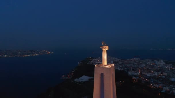 Noční dron létající kolem velké osvětlené sochy Ježíše na podstavci nad řekou. Letecký pohled na Kristovu svatyni v Almadě po západu slunce. Lisabon, hlavní město Portugalska. — Stock video
