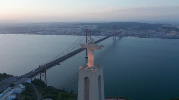 Vista traseira aérea da noite da estátua grande do Jesus no pedestal e na ponte longa sobre o rio. Drone voando em torno de Cristo, o Rei Santuário em Almada no tempo crepúsculo. Lisboa, capital de Portugal. — Vídeo de Stock