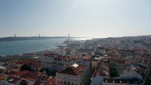 Panoramic aerial view of town with long cable-stayed bridge spanning Tagus river. Drone camera descending between buildings and gradually hiding view. Lisbon, capital of Portugal. — Stock Video
