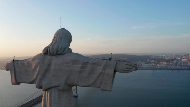 Een drone camera die rond het stenen Jezus standbeeld vliegt. Avond uitzicht op Christus de Koning Heiligdom in Almada over Tegus stroomt in het ondergaan van de zon. Lissabon, hoofdstad van Portugal. — Stockvideo