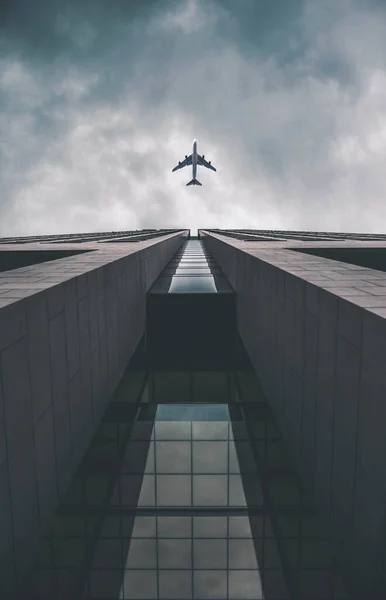 Low angle view of modern tall business building. Commercial jet plane flying over skyscraper. Against dramatic cloudy sky before storm. — Stock Photo, Image