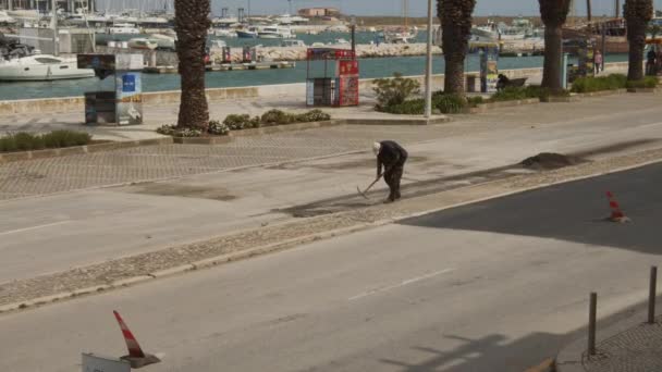 Bekerja sendirian menggali dengan tembilang. Mengambil lapisan yang rusak dari permukaan aspal di jalan sebelah marina. Jalan di Lagos, Portugal — Stok Video