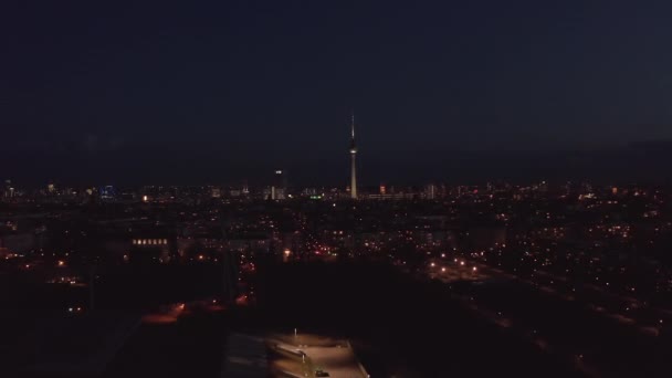 Vuelo sobre el barrio urbano por la noche. Vista aérea de calles y edificios con poca luz. Torre de televisión alta y delgada Fernsehturm con luces parpadeantes rojas. Berlín, Alemania. — Vídeos de Stock