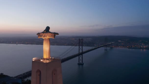 Órbita aérea ancha de gran monumento alto del Santuario de Cristo Rei con luces y puente rojo Ponte 25 de Abril a través del mar cerca de la costa de Lisboa, Portugal por la noche — Vídeos de Stock