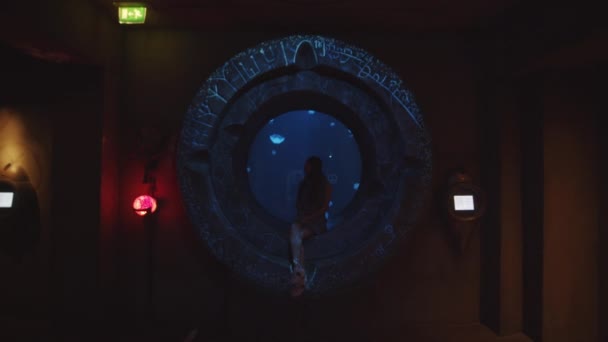 Langzame beweging van mooie jonge vrouw zittend op een cirkelvormige bandenstoel kijkend naar prachtige kwallen die zwemmen en bewegen op onderwaterachtergrond in het Dubai Aquarium — Stockvideo