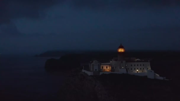 Static aerial view of Lagos lighthouse against cloudy dark blue sky at dusk, light flashing creates lens flare — Stock Video