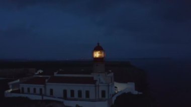 Portekiz 'in Lagos kasabasında alacakaranlıkta parlayan deniz feneri lambası, etrafta uçuşan hava aracı görüntüsü, parlayan mercek, alacakaranlık