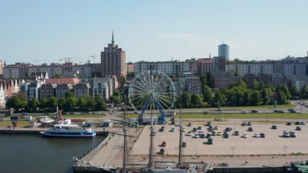 Slide and pan shot of waterfront with moored ships and Ferris wheel at large parking lot. Buildings in urban neighbourhood in background — 비디오