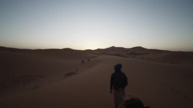 Önümüzde deve sürüşünden görüntüler var. Kefenli adam çölden geçen develerden oluşan kervana karşı korunuyor. Fas, Afrika