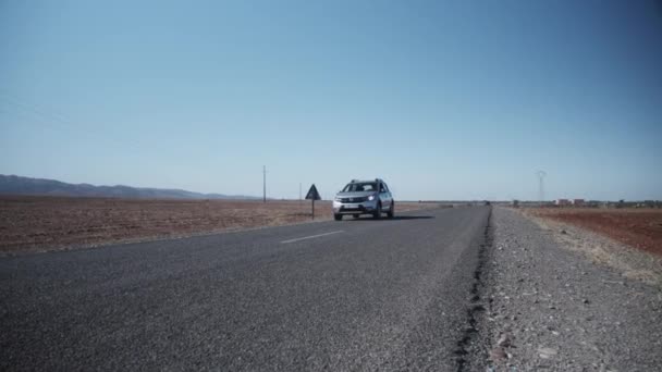 Ein Schwenk im Tiefflug zeigt einen silberfarbenen Geländewagen, der in trockener Landschaft mit Bergen in der Ferne auf der Straße fährt. Marokko, Afrika — Stockvideo