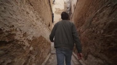 Yüksek binalar arasında dar bir sokakta yürüyen bir adamın arka görüntüsü. İhmal edilmiş ve otantik bir yere giren birinin düşük açılı görüntüsü. Fas, Afrika