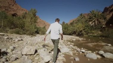 Nehir boyunca engebeli araziye giren bir adamın görüntülerini izliyorum. Taştan taşa zıplıyorlar. Turistlerin manzaranın tadını çıkardığı arka plan manzarası. Fas, Afrika