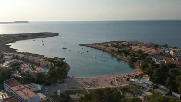 Vista panorámica aérea en cámara lenta de una multitud de personas irreconocibles relajándose y disfrutando en la playa rodeada de resort y mar con montaña durante la noche en Ibiza en España — Vídeos de Stock