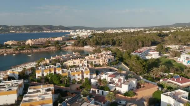 Vista circular aérea do telhado de belas casas perto da praia e da costa com barcos e iates estacionados rodeados por oceano e montanha em Ibiza, na Espanha, durante uma manhã cedo — Vídeo de Stock