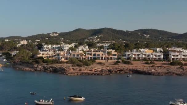 Vista aérea hacia adelante de los barcos estacionados en un mar silencioso y tranquilo con un joven parado en la orilla mirando a través de casa y complejo villa con vegetación en Ibiza en España — Vídeos de Stock