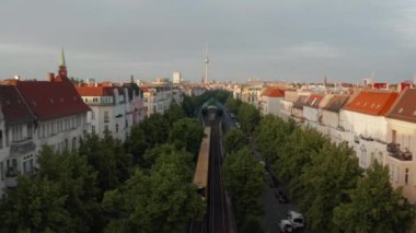 Geniş caddede ağaçlarla giden tarama trenini geriye doğru takip et. Etrafındaki binalar parlak sabah güneşiyle aydınlanıyor. Ufukta Fernsehturm TV kulesi var. Berlin, Almanya