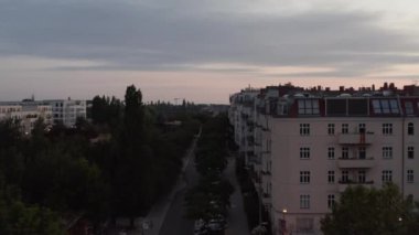 Forvetler caddenin üzerinde uçuyor ve şehirde yeşillik var. Tren yolu üzerindeki köprüde cadde devam ediyor. Alacakaranlık gökyüzü. Berlin, Almanya