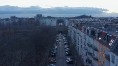 Berlin, Almanya 'da bulutlu bir sabahta, geleneksel binalar ve ağaçlarla çevrili caddenin bir tarafına park etmiş araçların tek yönde hareket ettiği ağır çekim görüntüsünü kurar.