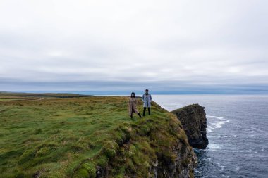İrlanda 'da Moher uçurumunun kenarında duran ceketli genç çift bulutlu gökyüzünün altında gün boyunca doğanın tadını çıkarıyor.