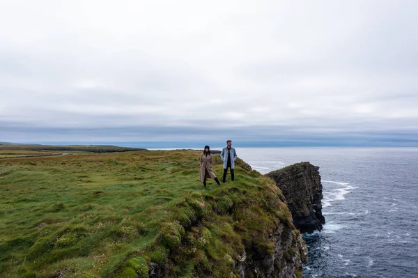 İrlanda 'da Moher uçurumunun kenarında duran ceketli genç adam ve kadın bulutlu gökyüzünün altında gün boyunca doğanın tadını çıkarıyor.