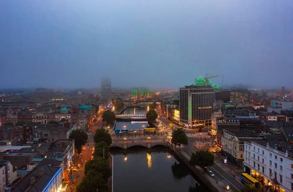 Dublin 'in sisli ufuk çizgisi ve caddenin iki tarafını birbirine bağlayan köprüyle akan nehir bulutlu bir gündoğumu sabahı aydınlatmalı ışıklarla çevrili binalar.