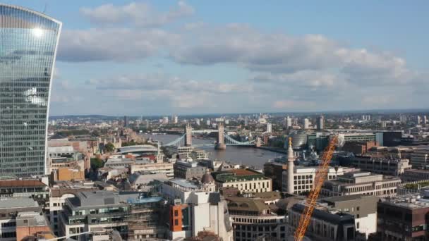 Vuela por encima del distrito de la ciudad. Fachada de vidrio en rascacielos moderno que refleja el cielo con nubes y sol brillante. Puente histórico de la torre sobre el río Támesis en el fondo. Londres, Reino Unido — Vídeo de stock