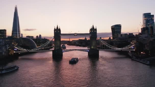 前方の水面の上にピンクの夕暮れの空を反映して飛ぶ。日没後にライトアップされたタワーブリッジの下に浮かぶ船。イギリスのロンドン — ストック動画
