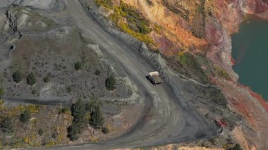 Engebeli bir arazideki madencilik operasyonunun hava görüntüsü, virajlı toprak bir yol ve çarpıcı kırmızı kaya oluşumları.