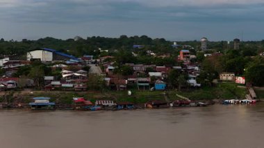 Hava perspektifi, bulutlu bir alacakaranlık sırasında Amazon yağmur ormanlarındaki bir nehrin çamurlu kıyılarında küçük bir köyü gösteriyor..