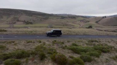 Siyah özelleştirilmiş 4x4 bir arabayı takip eden hava görüntüleri asfalt bir yolda engin, tepe kırsal bir arazide ilerliyor..