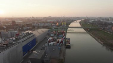Günbatımında nehir kanalı üzerinde bir konteyner terminali ve kargo gemisi bulunan Mannhees sanayi limanının havadan görüntüsü. Büyük depo ve şehir manzarası.