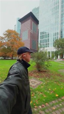 Bir adam, modern ofis binaları ve sonbahar yapraklarıyla çevrili bir şehirde parkta yürüyor..